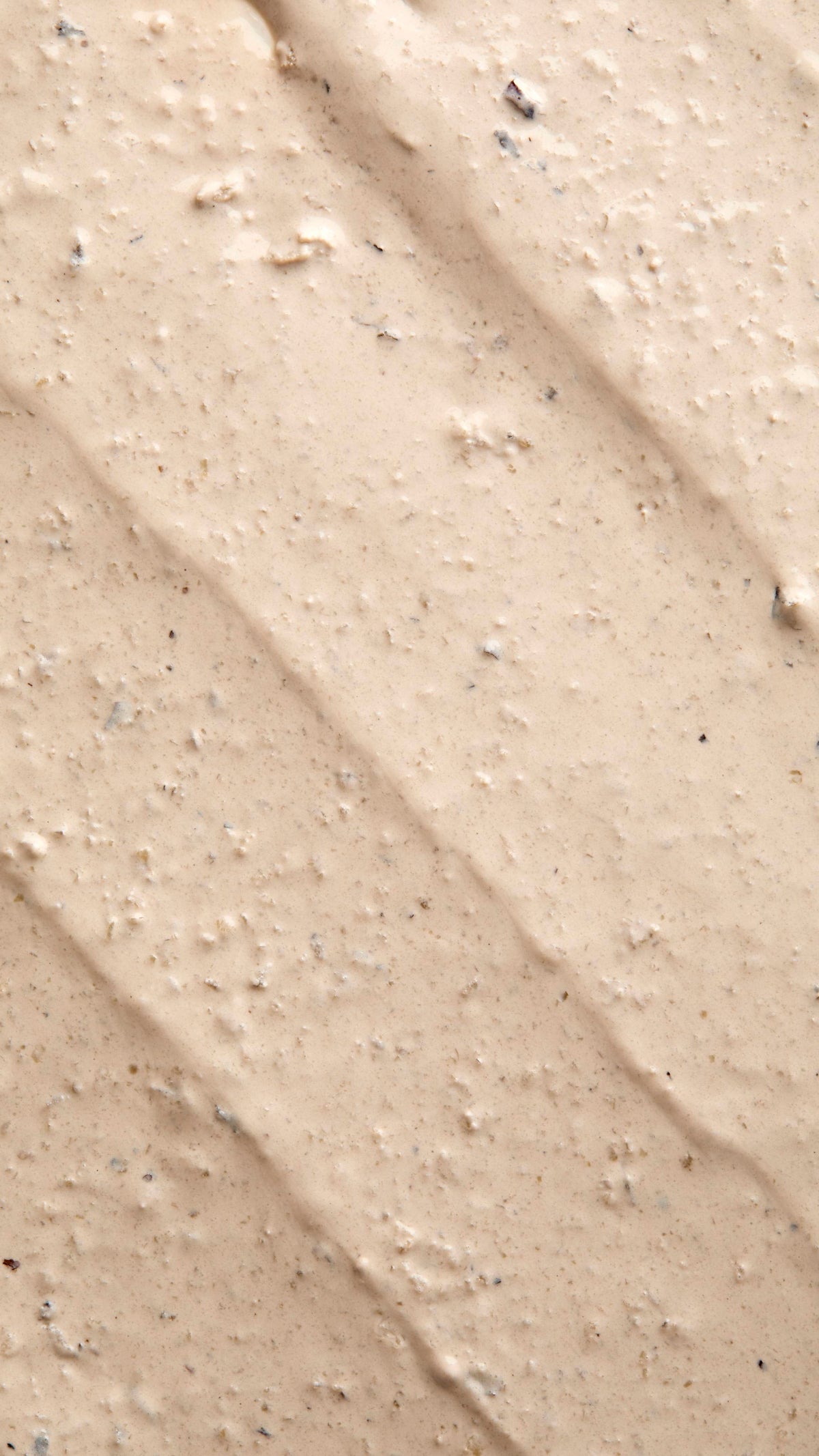 A close-up, textured shot of "Tiny Musicians Of The Forest, a creamy, beige-coloured face mask spread in thick, diagonal ridges. The surface is grainy and lumpy, containing small dark exfoliating particles and flecks.