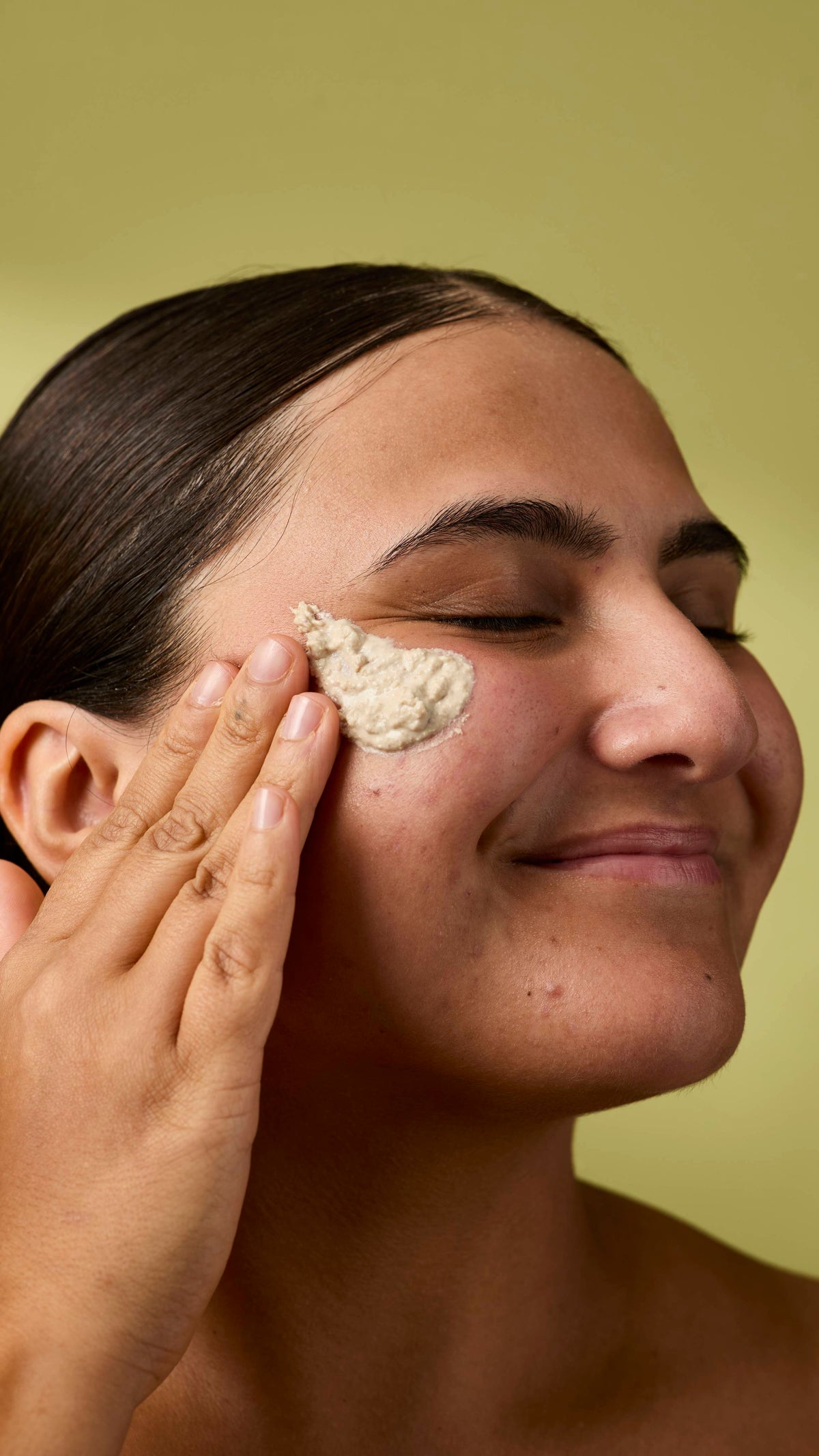 A close-up image of the model's face on an olive-green background. Their dark hair is tied back, and they are applying the textured Biomic facial cleanser to the skin on their cheek with their eyes closed.