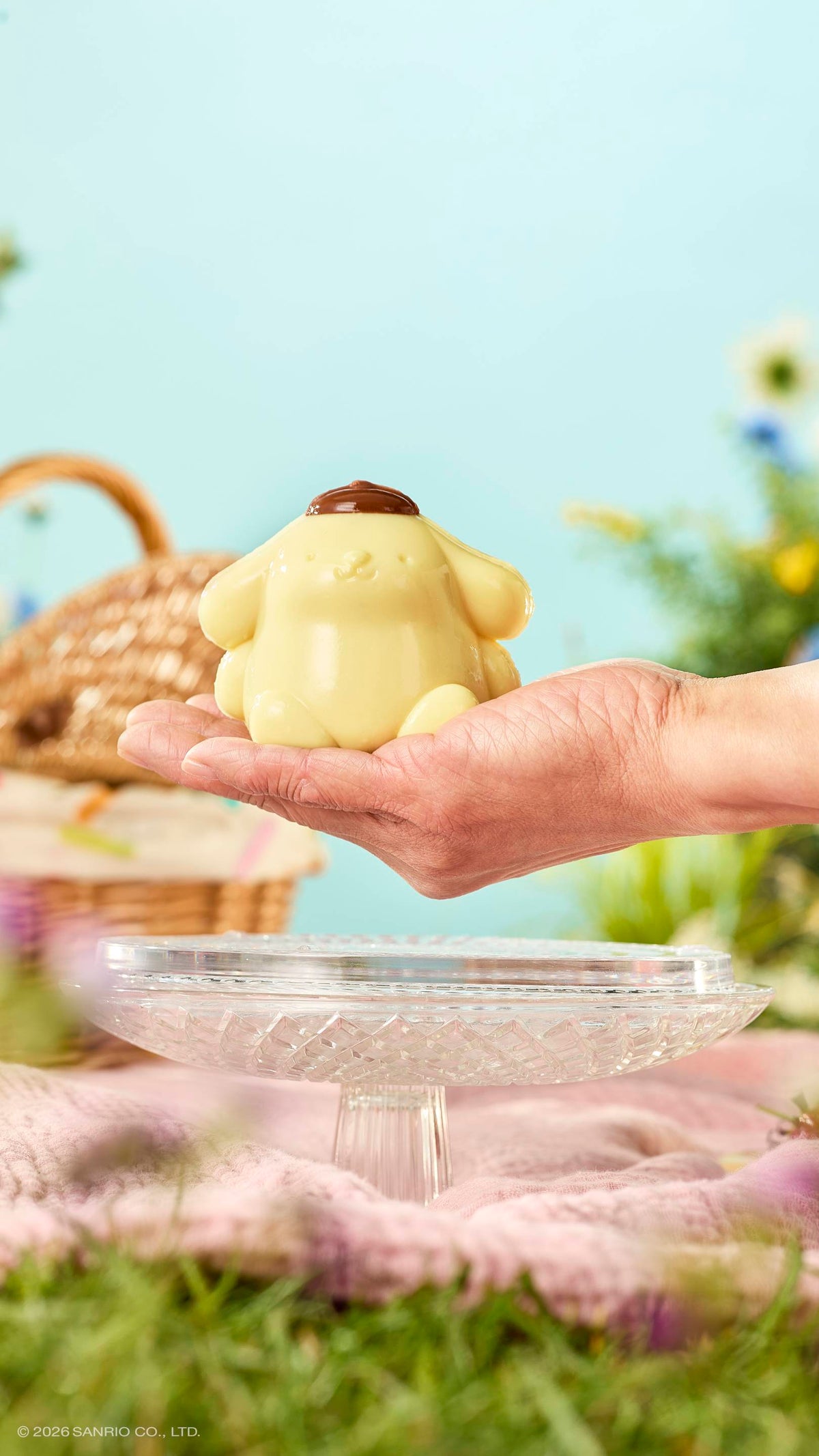 A hand holds a glossy, pale yellow "Pompompurin" shower jelly over a glass cake stand in a sunny meadow. The dog-like character wears a brown beret. A wicker basket and pink blanket sit on the grass under a blue sky.