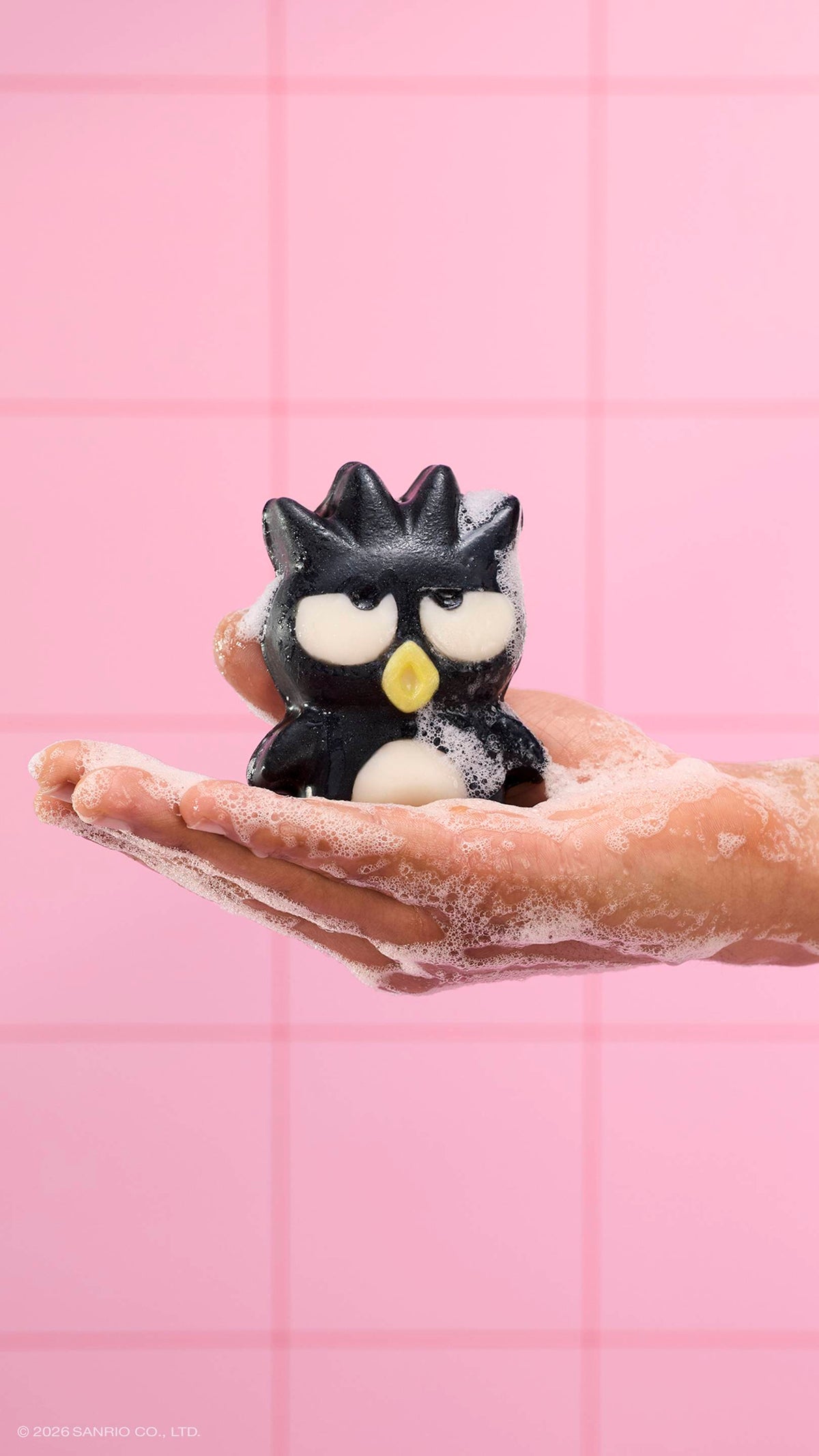 A soapy hand holds the black penguin-shaped "Bad Badtz-Maru" character soap, covered in white bubbles. The scene is set against a background of pink square tiles.