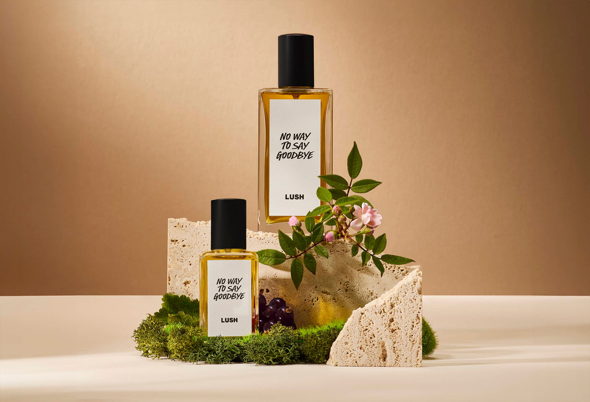 The image shows a soft, beige background. In the centre of focus are two bottles of two different sizes of perfume sitting on top of moss and rock with moss, blackcurrant and litsea cubeba flowers.