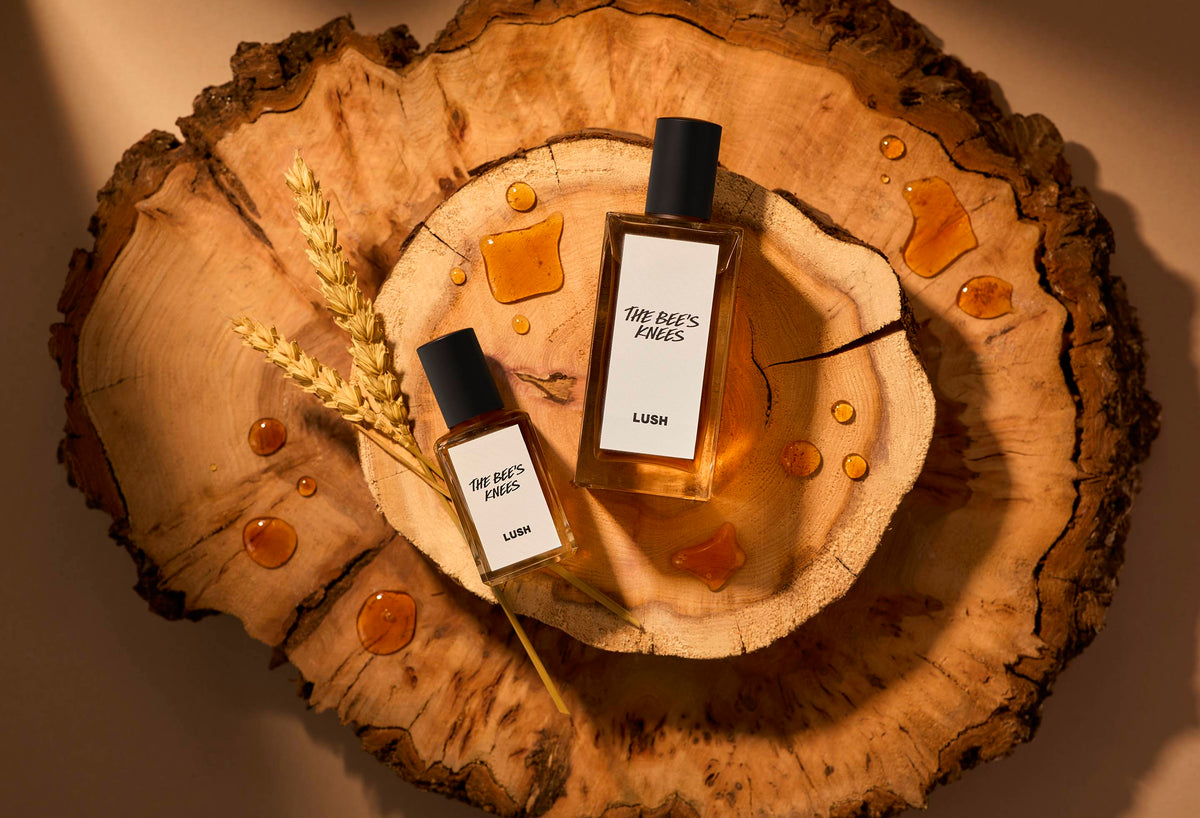 The image shows two stacked slices of wood. There is a warm light beaming over the two sizes of perfume bottles laid flat next to strands of wheat and dripped with splashes of honey.
